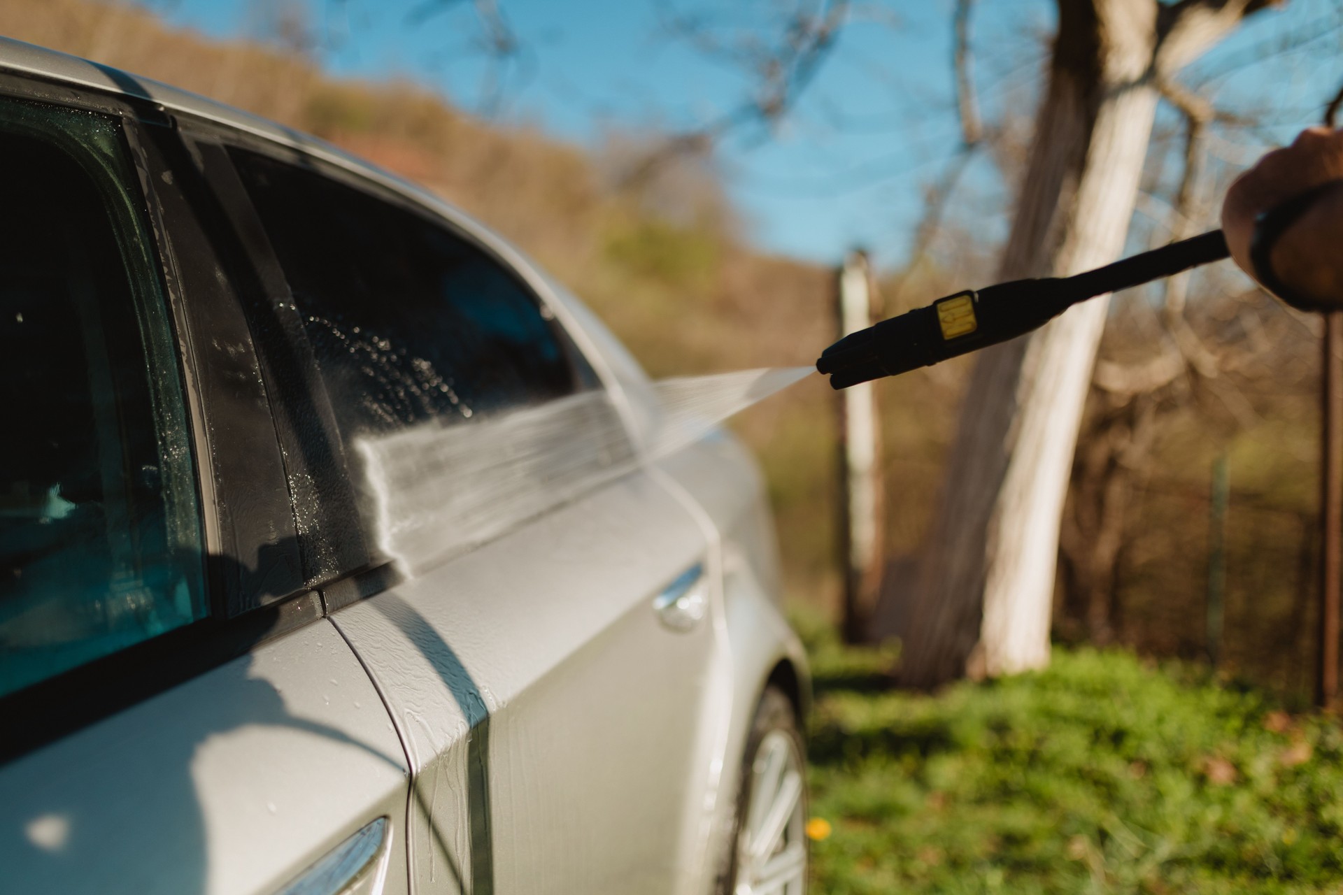 Man Using High-Pressure Washer to Clean Car Outdoors in Sunlight